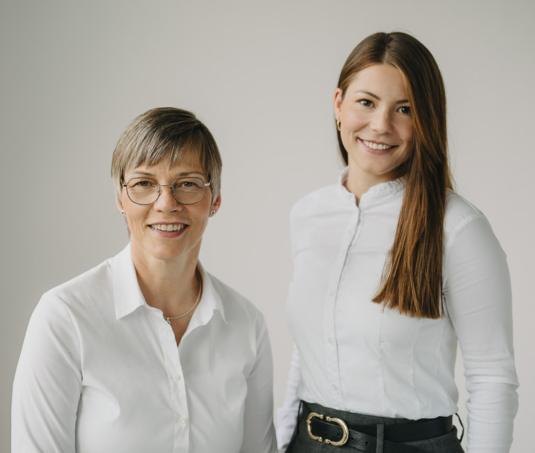 Zwei freundliche Frauen in weißer Bluse stehen vor neutralem Hintergrund und lächeln. Sie sind die Hauptansprechpartnerinnen für die Bestattungsvorsorge und beraten kompetent zu allen Fragen rund um das Thema.